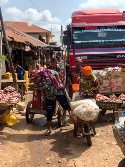 The image depicts a busy street scene in Ondo, Nigeria, at coordinates 7.251° N, 5.196° E. A woman in a blue and black patterned shirt and a red headscarf is pushing a wheelbarrow loaded with two large sacks of white powder. She is walking past a large red truck parked on the right side of the street. The truck is carrying a large load of goods, including several boxes labeled "Golden Morn" and "Danka Sulia." The woman is being followed by an older woman with a white headscarf and a green and brown dress. She is holding a bright orange shopping bag and a large white bag. Several other people can be seen in the background. The street is dusty and there are several buildings and stalls on both sides. There are several piles of onions on the ground, and a yellow bucket to the left of the wheelbarrow. A third woman is standing to the right of the frame, and she is partially obscured by the truck.