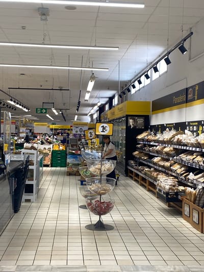 The image shows the interior of a supermarket in Bel Air, Spain. The perspective is from an aisle looking towards the front of the store, which is brightly lit by fluorescent and track lighting. The floor is tiled with large, square, off-white tiles. On the left, there are shelves with what appear to be frozen goods or storage units. Further down the aisle on the left, there are displays of fruits and vegetables, including oranges in green crates and watermelons. Signs for "SUPER ECONOMICO" are visible.
To the right, a large display of baked goods, identified by a "Panaderia" sign, stretches along the wall. The shelves are filled with various types of bread and pastries packaged in plastic bags. A woman in a uniform, likely an employee, is visible behind a counter in the middle of the store, interacting with products. In the foreground on the right, there are clear glass bowls displaying what look like small jars of preserves or condiments. Yellow signage with prices like "1€" is interspersed throughout the displays. The overall environment suggests a well-stocked grocery store.
