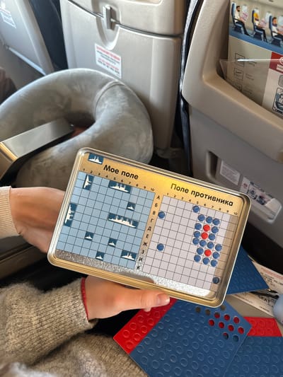 The scene depicts a passenger's view within an airplane cabin, identified as being in Aeroport, Russia. A person, wearing a light grey textured sweater with a red cuff partially visible, holds a metallic, rectangular board game. The game board is divided into two sections: "Мое поле" (My field) on the left, displaying multiple white ship silhouettes arranged on a light blue grid, and "Поле противника" (Opponent's field) on the right, showing a grid with numerous blue and several red circular pegs inserted. Both grids are labeled with numbers 1-10 and Russian letters A-K. Below the game board, several perforated plastic sheets, colored red and blue, are visible, along with a partially obscured document appearing to be a boarding pass or ticket stub with text including "SE ISSUED," "ODNOKINA POLINA," and "UAFAR08." In the background, a light grey neck pillow rests on an adjacent seat, and the grey seatbacks of an airplane are visible, one featuring a clear pocket containing an inflight safety card or magazine showing text in English and Russian, including "Taxing," "Take-off," and "Landing."