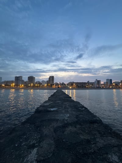 The image, when correctly oriented, depicts a serene coastal scene of a city at either dusk or dawn. The upper half of the frame is dominated by a vast sky, transitioning from a deeper blue at the top to lighter, more subdued hues of purple and pink closer to the horizon, characteristic of twilight. Wispy, elongated clouds are scattered across the sky, adding texture and depth.

Along the horizon, a city skyline stretches across the mid-ground. Numerous multi-story buildings are visible, many with their interior or exterior lights illuminated, casting warm, yellowish glows. These city lights create striking, elongated reflections on the calm surface of the water, which occupies the left side of the lower frame. The water shows gentle ripples, indicating a light breeze or slight current, and effectively mirrors the sky and urban illumination.

A dark, rugged concrete pier or breakwater extends diagonally from the lower right foreground towards the center of the image, gradually narrowing into the distance. This structure provides a strong visual anchor and separates the open water from the more developed coastal line. No individuals are visible, contributing to the quiet and contemplative mood of the scene. The image captures a peaceful moment along the waterfront of a coastal city, confirmed to be Vinaròs, Spain. There is no visible text in the media file.