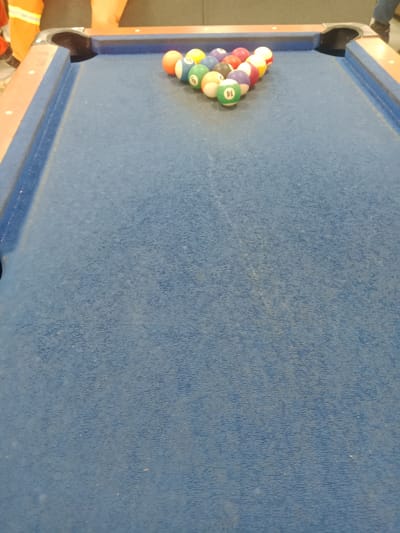 A rack of pool balls sits on a blue felt billiard table in an indoor setting. The balls are arranged in a triangular formation, with the cue ball at the apex. Some of the balls are numbered and colored, while others are solid colors. In the background, two people are partially visible, wearing orange vests. The lighting suggests it is daytime. The scene appears to be a casual or recreational environment, possibly a game room or a bar. No specific location within Kwale, Nigeria is discernible from the image itself.