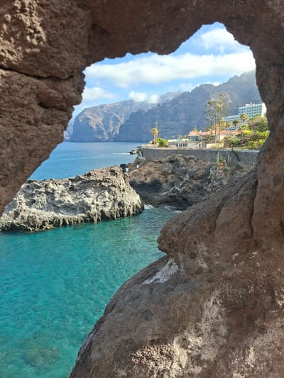 The image depicts a coastal scene viewed through a natural rock archway. In the foreground, rough, textured brown rocks frame the view. Beyond the rocks, a body of clear, turquoise water stretches out, meeting a rocky coastline. A large, jagged rock formation juts out from the sea on the left.
Further inland, a paved pathway with railings and steps winds along the rocky cliff face. On the upper level of the cliff, several buildings are visible, including a white, multi-story hotel with many windows and what appears to be a resort complex with red-tiled roofs and balconies. Palm trees and other lush vegetation are scattered throughout the landscape.
In the background, imposing, dark, and rugged mountains rise sharply, their peaks partially obscured by a hazy sky with scattered white clouds. The scene suggests a scenic coastal resort area, likely in a volcanic region given the rock formations. The location is identified as Santiago del Teide, Spain, suggesting this is a view of the Atlantic coast of Tenerife, known for its dramatic cliffs and coastal resorts. There are no people visible in the image.