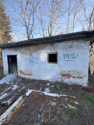 An abandoned, single-story, rectangular structure with heavily degraded white plaster walls, revealing red brickwork, stands in the city of Stoianovo, Bulgaria. The building features an open doorway on the left and an empty window opening on the right, both devoid of frames or coverings, leading to dark interiors. Blue spray-painted graffiti is visible between the door and window, and "1912!" in green spray paint is present to the right of the window. The structure has a dark, flat concrete roof. The surrounding ground is covered with dry brush, bare earth, sparse green vegetation, and scattered debris including white panels with exposed insulation. Patches of snow are present near the doorway. Deciduous trees with bare branches are visible in the background against a clear blue sky. No individuals or active elements are present in the scene.