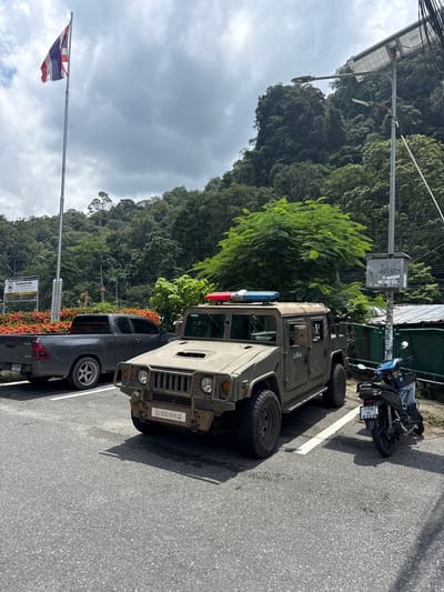 The image depicts an outdoor scene in Tambon Wang Prachan, Thailand, featuring parked vehicles and a lush, forested hillside. A tan-colored military-style vehicle, possibly a Humvee, is parked prominently in the foreground, equipped with red and blue emergency lights on its roof. To its left, a dark gray pickup truck is parked. A black motorcycle with a light blue case is parked to the right of the military vehicle. A Thai flag flies on a tall pole to the left of the frame. In the background, a steep, tree-covered mountain rises under a cloudy sky. The ground is paved asphalt, with white parking lines visible. Several solar-powered streetlights are also present. A sign with Thai text and a prohibition symbol is visible near the pickup truck. The overall environment suggests a location near a natural border or a checkpoint, given the presence of the military vehicle and the Thai flag.