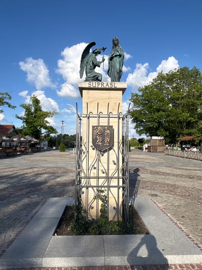 The image shows a bronze statue of an angel kneeling before a woman, with the word "SUPRASL" inscribed above them on a stone monument. The monument is surrounded by a decorative metal fence with a coat of arms featuring two winged figures. The scene is set in a public square paved with cobblestones, with trees and buildings in the background. The sky is blue with white clouds. There are some people visible in the distance, as well as outdoor seating areas with umbrellas. The statue appears to be a significant landmark in the town of Ogrodniczki, Poland, possibly commemorating a religious or historical event.