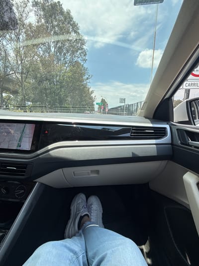 A view from inside a car, likely in Sanctorum, Mexico, given the road sign partially visible. The view through the windshield shows trees and a blue sky with clouds. Inside the car, the dashboard is visible, including a digital screen displaying a map. A person wearing light blue jeans and grey sneakers is sitting in the passenger seat. The overall environment suggests daytime travel on a road with trees nearby.