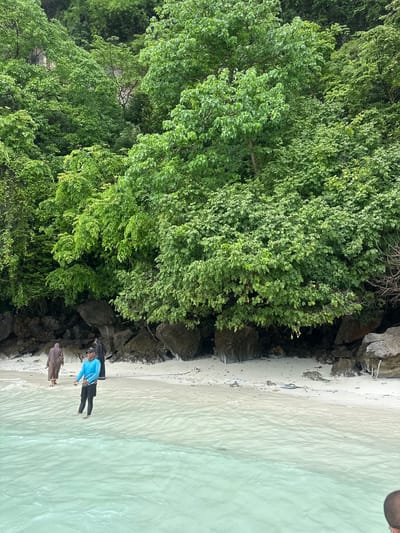 This is a medium shot of a beach with lush green trees in the background. The water is clear and turquoise. In the foreground, a man in a blue long-sleeved shirt and black shorts is standing in the water, holding a rope. Behind him, two people are walking on the beach; one in a brown robe and the other in a black garment. The beach is sandy and rocky with some debris scattered on it. The image appears to have been taken from a boat or a low vantage point.
