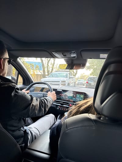 Captured from a backseat perspective inside a car, the driver is visible holding the steering wheel of a Kia. A digital display screen is mounted on the dashboard. Outside the car, a line of vehicles and trees can be seen. The interior includes dark gray upholstery with headrests, a sun visor, and a rearview mirror. The light suggests it's daytime.