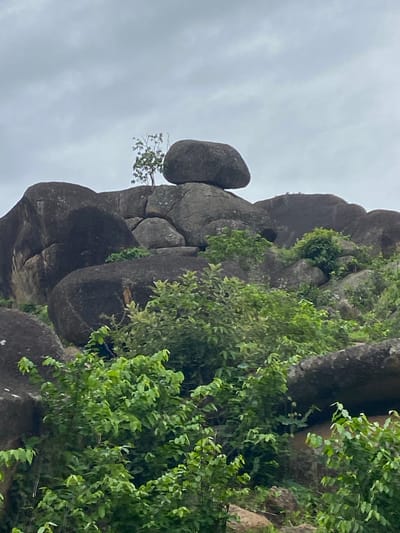 The image shows a cluster of large, smooth, dark gray rocks stacked precariously on top of each other. A small tree grows out from the rocks. Green foliage is visible on the side of the rock formation. The sky above is overcast and gray. The rock formation is located in Ganawuri, Nigeria.