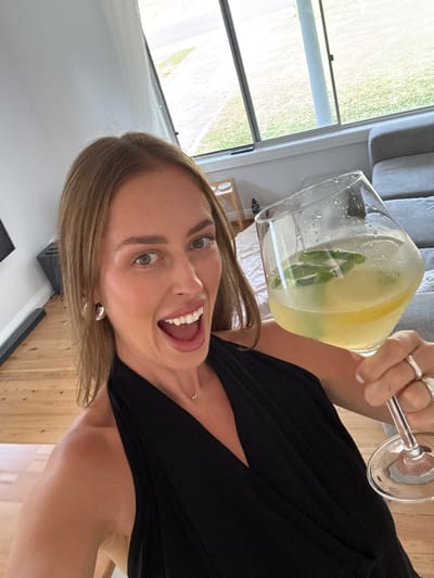 A woman holds a large glass of liquid containing ice and a green sprig. She is smiling widely, her mouth open, with light brown hair and a black top. Visible behind her is a window with a view of greenery and a gray couch. Her rings are visible on the hand holding the glass. She is indoors, likely in a home setting. The image is angled as if taken in selfie mode.