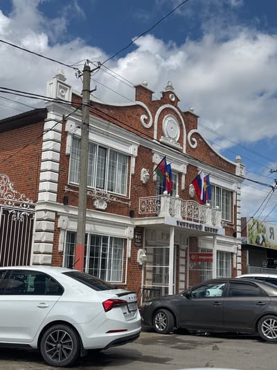 The image shows a three-story brick building in Yelizavetinskaya, Russia, with distinctive Dutch gable architecture. The building has white trim and decorative wrought iron elements. Two cars are parked in front of the building: a white MG sedan and a dark gray sedan. The building flies the flags of Adygea. Power lines and a utility pole are visible in the foreground. The sky is partly cloudy.