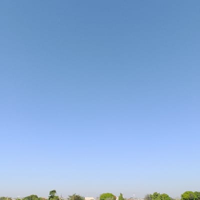 The image presents an outdoor scene dominated by a clear, light blue sky, which occupies approximately 90% of the frame. Along the lower horizon, the tops of green trees are visible, interspersed with the upper portions and rooftops of various distant buildings. These structures appear to be residential, featuring light-colored walls and some reddish-brown tiled roofs. No individuals, specific subjects, actions, or interactions are discernible within the visible landscape. The environment suggests a bright, sunny day over a populated or semi-rural area within Estômbar e Parchal, Portugal.