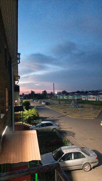 This is a vertical, slightly grainy photograph taken from an elevated vantage point, looking out over a residential area at dusk. The sky is a gradient of deep blue at the top, transitioning to softer blues and pinks near the horizon, suggesting the sun has recently set. Wispy clouds are scattered across the sky.
In the foreground to the left, the brick facade of a building is visible, with a balcony railing and a section of a wooden roof. Below this, a street curves through the scene, lined with parked cars. Several silver sedans are parked along the street and in a lot.
Further into the scene, a row of low-rise houses with light-colored walls and dark roofs stretches across the middle ground. They appear to be part of a residential complex. In front of these houses, a small playground with a slide and swing set is visible. Beyond the houses, trees and more buildings are sparsely distributed, and a tall industrial chimney with a light on at its peak stands prominently in the distance. The ground is covered with dry grass and patches of dirt, indicating a potentially dry climate or season.
The overall impression is one of a quiet evening in a suburban or semi-rural setting. The time of day is clearly dusk, with the fading light casting long shadows and creating a serene atmosphere. There is no visible text in the image.