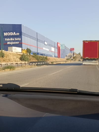 A long, blue building with "MODA Life" and "Fabrika Satış Mağazası" written in white lettering dominates the left side of the frame. The building appears to be a large retail store, possibly a factory outlet given the text. Adjacent to it are smaller structures and some yellow and white shipping containers. The building is situated on a hillside covered in dry grass and scattered bushes and trees. A highway runs in front of the building, with a barrier separating it from the embankment.

On the right side of the highway, a red semi-trailer truck is visible, its back doors closed. It has a license plate that reads "07 ABS 465". The truck appears to be moving along the highway, as is the perspective of the viewer, who is inside a vehicle.

The scene is set under a clear, bright blue sky, suggesting a sunny day. The sunlight casts shadows, indicating it is likely daytime, possibly late afternoon due to the angle of the sun. The landscape is arid, typical of a dry climate. The overall impression is one of commercial activity along a transportation route in a somewhat rural or semi-urban setting. The text on the building and the truck's license plate suggest a location in Turkey. The text on the truck, "Mint Stouligionen Af Diterim", is likely a slogan or company name in Turkish. The scene appears calm and uneventful, focusing on the infrastructure and commercial presence.