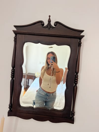 A young woman with blonde hair is depicted in a selfie, reflected in an ornate, dark brown wooden mirror. She wears a light-colored tank top and light blue denim jeans, holding a dark-colored smartphone in her right hand at chest level, with a grey lanyard attached. Her left hand is partially visible at her side, adorned with a black bracelet on her wrist. The mirror frame features a carved finial and scrollwork at the top, turned posts along the sides, and a scalloped bottom edge. In the reflection behind the woman, a bedroom environment is visible. This includes plain white walls, a bed with a light-colored duvet, and a headboard or pillows with yellow and blue stripes. To the left of the bed, a dark wooden nightstand with a single drawer and turned legs is present. Two square, light-colored fixtures, possibly outlets or switches, are visible on the white wall. A light-colored candle is partially visible in the bottom left foreground, outside the mirror's reflection. The scene is located in Oliveira de Frades, Portugal.