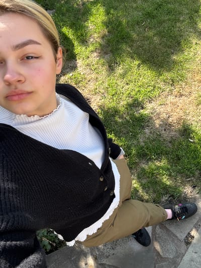 The image shows a young woman wearing a black cardigan over a white shirt and khaki pants. She is standing on a paved area next to a patch of grass. The grass is green with brown patches. The woman's face is visible in the photo, and she appears to be looking at the camera. The woman is wearing black shoes. The photo appears to be taken in a park in Crail, United Kingdom. The coordinates provided correspond to a location near the shoreline of the town of Crail, a seaside town in Fife, Scotland.