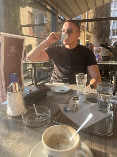 The image depicts an outdoor or semi-enclosed cafe setting at "Ulrich Das Stadtcafé" located in Krems an der Donau, Austria.
A male individual, appearing to be in his 20s or 30s, is seated at a light-colored, textured table. He has short, dark hair and is wearing rectangular-frame sunglasses and a black t-shirt with a dark emblem on the left chest. Red string bracelets are visible on his left wrist. He is actively drinking from a small white espresso cup held in his right hand, with the cup at his lips.
The table in front of him holds various items:
* In the immediate foreground, a white ceramic espresso cup, largely empty with coffee residue, contains a silver spoon and rests on a matching saucer.
* To the left, an open glass ashtray contains three cigarette butts and ash, with one cigarette standing upright.
* Further left, a silver-capped, fluted salt or sugar shaker is positioned next to a menu board for "Ulrich DAS STADTCÁFE."
* Behind the ashtray, a dark-colored smartphone lies face up next to a black eyewear case and a blue cigarette lighter.
* Towards the right side of the table, a small white ceramic creamer or milk jug is present.
* A small white plate with a silver spoon rests near the creamer.
* Two tall, clear glasses filled with water are visible; one is directly in front of the subject, and the other is on a rectangular silver tray.
* The silver tray also holds a white paper napkin covering several white sugar packets (labeled "Lavazza") and other small sachets. A pack of cigarettes is partially visible behind the second water glass.
In the background, visible through a glass enclosure, other blurred cafe patrons are seated at separate tables. One female figure with light hair is partially discernible directly behind the main subject. Strong natural sunlight illuminates the scene, casting reflections on the glass panels and highlighting the table surface. Architectural elements of surrounding buildings are visible in the background reflections and directly through the glass.