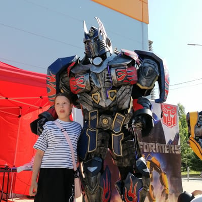 The image depicts an outdoor scene in Трубачеевка, Russia, featuring a young girl standing next to a person in an elaborate Optimus Prime costume.
The girl, positioned on the left, has light brown hair pulled back. She wears a white t-shirt with thin black horizontal stripes, a dark-colored skirt, and a light pink cross-body bag with a wide strap. Her mouth is slightly open, and she looks towards the left of the frame.
To her right, a tall, detailed costume of Optimus Prime stands prominently. The costume is multifaceted, appearing worn with various textures, and features a metallic-like finish in shades of silver, dark blue, red, and black, with gold accents. Red flame decals are visible on parts of the costume, particularly on the chest and legs. Small blue LED lights are integrated into the costume, notably on the head and torso.
In the background to the left, a bright red pop-up canopy tent is visible, under which a table holds various electronic devices and cables. To the right of the Optimus Prime costume, a large vertical banner is displayed. The banner features the red Autobot symbol at the top and the word "TRANSFORMERS" in metallic silver lettering, with an image of a yellow robot, identified as Bumblebee, on its lower half. Further to the right, a partial view of another yellow robot costume, likely Bumblebee, is visible.
The far background includes a modern building with orange and grey paneling under a clear, light blue sky. Thin black power lines traverse the upper portion of the sky. A distant street lamp and green foliage are also visible on the right. The scene is brightly lit by natural daylight.