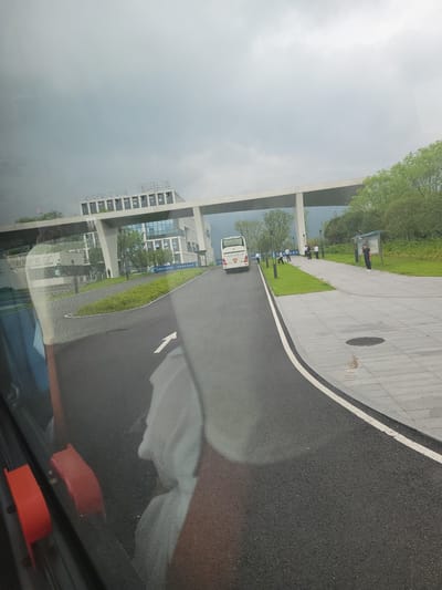 The image captures a cloudy day scene in the city of Yufengshan, China. In the foreground, a paved road curves to the right, marked with a white line and a white arrow indicating direction. To the left of the road, a green grassy area is bordered by a concrete structure. Beyond this, a modern building complex is visible, with large glass windows and a white overpass connecting sections of the building. A white bus with a license plate containing "0 E5337" is parked on the road. Several individuals, appearing to be men, are standing near the bus and on a paved walkway. The walkway is adjacent to a grassy area with trees and vegetation. In the background, the sky is overcast, and a line of trees is visible on a hillside. The reflection of the viewer and the interior of a vehicle are also visible in the lower left portion of the image.