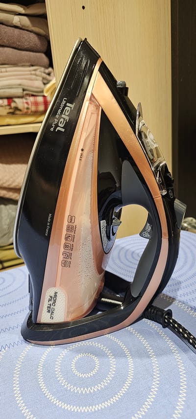 The image shows a close-up, side view of a Tefal Ultimate Pure steam iron, predominantly black with rose gold accents, resting on a blue ironing board cover with a white spiral pattern. The iron is positioned to the right of the frame, with its handle facing the viewer. The water reservoir, a translucent pinkish material, is partially visible and contains a small amount of water with some bubbles. The word "Tefal" is clearly visible on the top of the iron, along with "Ultimate Pure." Below this, text indicating "Made in France" is present on the left side. A "MICRO CALC FILTER" label is visible on the base of the iron. On the rose gold section of the iron, there are several icons, including "3200W," a representation of steam, "auto-steam," "anti-drip," and "anti-calc." The iron is plugged in, with a black and white patterned power cord extending to the right and slightly out of frame. In the background, a wooden shelf unit with stacked folded fabrics is visible. The scene is indoors, likely in a home setting. There are no people or animals present in the image. The lighting appears to be artificial, illuminating the iron and its surroundings. The location is not specified in the image.