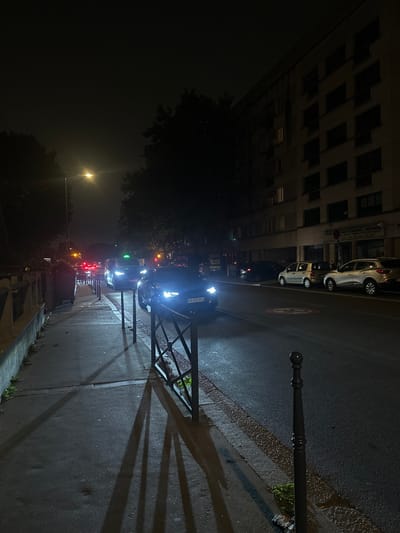 This image depicts a nighttime street scene in Colombes, France. The perspective is from a paved sidewalk, looking down a street with vehicles and buildings.
In the foreground, a dark-colored metal barrier, approximately waist-height, separates the sidewalk from the asphalt road. This barrier casts elongated, sharp shadows onto the sidewalk due to an unseen light source to the right, suggesting strong artificial illumination. A dark, ornate metal bollard stands in the lower right corner, marking the edge of the sidewalk. The sidewalk itself is concrete, displaying minor surface irregularities and patches of vegetation near the curb and barrier. A concrete retaining wall or ledge is visible further down the sidewalk on the left, with a dark, cylindrical object (possibly a trash receptacle) behind it.
The asphalt road, which appears damp or reflective in sections, occupies the central and right portions of the image. A white circular road marking with a red border, indicative of a speed limit (likely 30 km/h), is visible on the road surface in the mid-right. Multiple vehicles are present on the road. In the immediate mid-ground, a dark-colored sedan, possibly a Renault Talisman, with bright LED headlights illuminated, faces away from the camera. Its license plate is partially visible, starting with "GH-890-". Behind this sedan, a dark van or large car, identified as a taxi by a green illuminated sign on its roof, also has its headlights on. Further down the street, red taillights from additional vehicles are visible in the distance, indicating traffic. On the right side of the road, several cars are parked parallel to the curb, including a light-colored compact car (possibly a Fiat 500) and a silver SUV (possibly a Renault Captur or Arkana).
On the left side of the street, a tall streetlamp with a bright, yellow-orange light source illuminates parts of the scene. Dark, dense tree foliage is prominent above the street, partially obscuring the dark, featureless night sky.
On the right side of the image, running parallel to the street, is a multi-story building with a light-colored facade. This building features multiple rectangular windows, most of which are dark, but several display interior illumination on different floors. At ground level, a commercial sign with French text is partially legible, indicating "TRAVAUX GENERAUX DEMENAGEMENT" (General Works Moving) and a phone number. No individuals are visible in the captured scene.