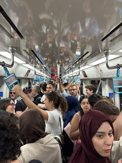 The image shows a crowded subway car in Zeytinburnu, Turkey. Many people are standing, holding onto overhead rails. There is a woman wearing a dark red hijab in the foreground, looking towards the camera. Other people are in close proximity, and some are holding onto blue grab handles. The subway car interior appears clean and modern.