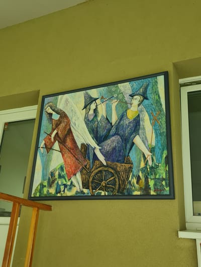 A painting is displayed on an olive-green textured wall. The painting, framed in black, depicts three figures. Two figures, dressed in pointed hats and dark robes, sit in a wooden cart. One figure plays a flute, blowing bubbles into the air. The third figure, wearing a red garment, stands next to the cart, holding two sticks. The background of the painting features abstract green and blue elements suggesting foliage or trees. The painting is positioned between two white window frames. To the left of the painting, a light brown wooden railing is partially visible. The setting appears to be an outdoor, possibly covered, area. The signature on the painting indicates it is by "SINTAR, 2002." This artwork is located on the exterior wall of the Molėtai Museum of Ethnography and Art in Molėtai, Lithuania.