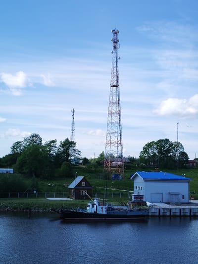 The image shows a waterfront scene in Ivanov Bor, Russia. A dark-colored tugboat is docked beside a light gray building with a blue roof. Behind the building is a grassy area with trees, and a smaller wooden building is visible. Dominating the background are two large communication towers, the taller one painted with red and blue sections. The sky is blue with scattered white clouds. The water is dark and reflects the surrounding scene.
