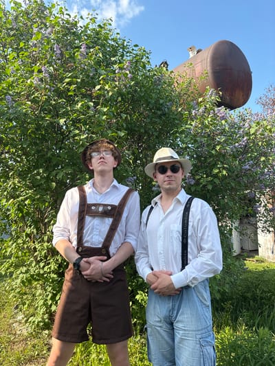 Two young men are standing in an outdoor setting amidst greenery and flowering bushes. The man on the left wears a white button-down shirt, light blue jeans, black suspenders, a light-colored hat, and sunglasses. The suspenders have "Columbia" printed on them. The man on the right wears a white button-down shirt, brown lederhosen, and suspenders with traditional decorative elements. He is wearing glasses and a brown hat. Their hands are clasped together. Behind them is dense foliage with lilac-colored flowers. To the left, a large rusty tank is visible against a blue sky. A small building is partially visible behind the foliage. The location is Пальники, Russia.