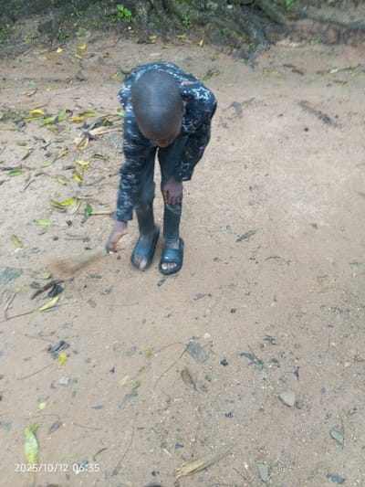 The scene, located in Kamantan, Nigeria, was recorded on 2025/10/12 at 06:35. It depicts a young individual, likely a child, viewed from an elevated perspective, bent forward and looking down at the ground. The person has short, dark hair and wears a dark, long-sleeved shirt with a scattered white star pattern, dark trousers, and dark open-toed sandals. The trousers, knees, and feet exhibit visible dirt. The individual's left hand is positioned near the ground, interacting with a small, elongated object, possibly a twig, which appears to be brushing or disturbing the sandy surface, evidenced by a slight plume of dust. The ground is composed of light brown sandy dirt, sparsely covered with small stones, dried leaves, and dark organic debris. Exposed tree roots and minimal green foliage are visible in the upper left background, indicating an outdoor, unpaved environment.