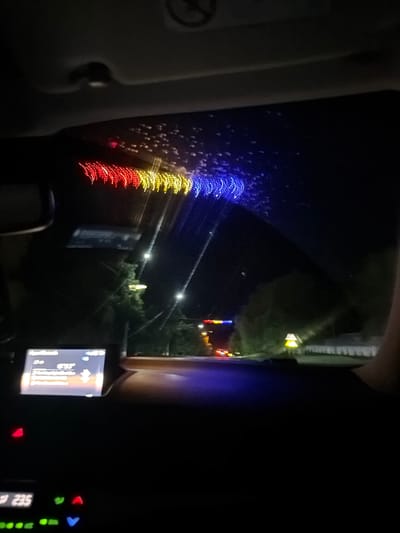 A car dashboard is visible in the foreground, with a screen displaying what appears to be navigation information. A digital display shows "235" and other illuminated buttons. The view through the windshield reveals a nighttime street scene. A string of red, yellow, and blue lights is draped across the top of the windshield, creating a bokeh effect. Outside the car, streetlights illuminate trees and a road. In the distance, traffic lights and other vehicle lights are visible. The location is specified as Chetroșica Veche, Moldova.