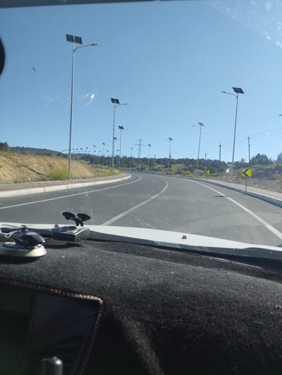 This is a photograph taken from the interior of a vehicle looking out at a road on a clear, sunny day. The road is paved and appears to be a highway or a major road, curving to the right in the distance. On either side of the road, there are streetlights with solar panels on top, indicating a focus on sustainable energy. The landscape alongside the road is hilly and covered with dry vegetation and some scattered trees. There are also power lines visible in the background, suggesting the presence of an electrical grid. Yellow directional signs are posted along the roadside, indicating curves in the road. The foreground of the image shows the dashboard of the car, with two objects mounted on it. One appears to be a suction cup mount, possibly for a phone or GPS device, and the other is a small black electronic device. The sky is a bright, clear blue with a few wispy clouds. The image does not depict any people or specific city landmarks that would definitively place it in Myk, Uzbekistan. However, the presence of solar-powered streetlights and the general arid landscape could be consistent with regions in Central Asia.