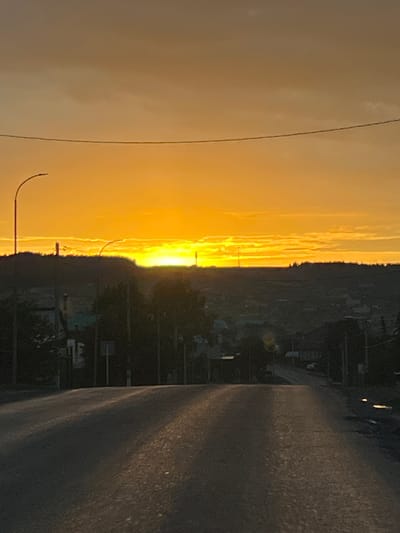 This is a medium-shot, vertical, outdoor, daytime photograph taken at dusk. The scene depicts a road in a rural or suburban area with a sunset in the background. The sky is a vibrant orange and yellow, with some clouds visible. A silhouette of a hill or treeline separates the sky from the landscape below. On the hill, there are several structures that appear to be buildings and possibly communication towers. The road in the foreground is paved and appears to be wet, with reflections of light on its surface. The overall mood is serene and peaceful, possibly indicating the end of the day.