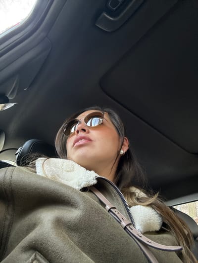 A woman wearing sunglasses and a jacket with a fur collar sits in a car, looking slightly down at the camera. The car's interior, including the sunroof and seats, is visible in the background. She has long, dark hair and is wearing a pearl earring. The jacket is a shade of green or brown, with light-colored straps or accents visible.