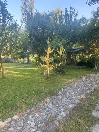 The image depicts a paved stone pathway leading through a lush green lawn dotted with trees. In the background, a small gazebo with a metal roof and green railings is visible, surrounded by dense foliage. The scene appears to be a park or garden area. The lighting suggests it is either early morning or late afternoon, with sunlight casting long shadows across the grass. The location is specified as Artavaz, Armenia, implying an outdoor recreational setting within the city.