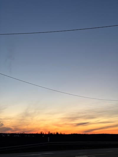 The image is an upward-angled landscape shot of the sky, likely taken near Kuregovo, Russia. Two power lines diagonally traverse the left side of the frame. The sky transitions from a deep blue on the left to a sunset of orange and yellow hues on the right. A dark silhouette of trees and possibly buildings sits along the right edge, alongside a road or barrier. The photo appears to be taken on or near a road with roadside barrier.