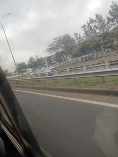This image captures a roadside scene in Gatongora ward, Kenya, viewed from within a moving vehicle. The foreground features the dark, angled interior structure of a vehicle's window frame. Immediately to the right, a dark asphalt road surface with a solid white line on its outer edge is visible. A metal guardrail extends horizontally across the midground, separating the immediate road from a grassy median strip. Beyond the guardrail, another multi-lane asphalt road carries traffic. Vehicles observed on this road include a light blue sedan, a light-colored bus with illuminated yellow lights, a white van, and a white car further down the road. A tall streetlamp with a single luminaire stands on the far left. The background reveals a row of trees with varying foliage, including some feathery varieties, fronting a series of multi-story buildings with light-colored facades. The sky is uniformly overcast, indicating daytime conditions.