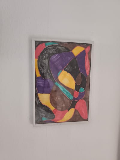 The image shows a framed, abstract painting hanging on a white, textured wall. The painting itself is composed of various geometric and organic shapes filled with bold colors: purple, black, yellow, teal, red, and brown. The shapes are outlined with black and appear to be drawn with markers or colored pencils. The composition is dynamic and somewhat chaotic, with interlocking shapes that create a sense of movement. There is no discernible subject matter like people or a specific scene, and no visible text is present. The overall impression is that of a piece of modern art. The setting appears to be indoors, likely a home or gallery, given the plain white wall and the way the artwork is displayed. The lighting is neutral, suggesting it could be daytime or artificial light. The image is taken from a slightly angled perspective, with the bottom edge of the painting slightly closer to the viewer than the top.