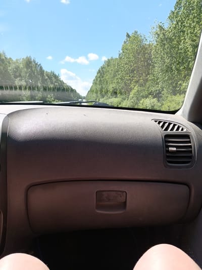 The image depicts the interior of a car, specifically the dashboard, with a view through the windshield. Outside, the scene shows a road flanked by dense green trees under a partly cloudy blue sky. A concrete barrier runs along the left side of the road.

The dashboard is dark gray and textured, featuring an air vent on the right side and a glove compartment below. A portion of the driver's legs is visible at the bottom of the frame.

The location is inferred to be near Uspenskii, Russia, based on the user's prompt, but there is no corroborating visual information within the image to confirm this location. The photo focuses primarily on the car interior and the immediate outdoor environment visible through the windshield.