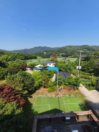 The image depicts a rural landscape in Seorak-myeon, South Korea, on a clear, sunny day. In the foreground, a wooden deck with a table and chairs overlooks a small, artificial turf court marked with white lines, resembling a mini-tennis or badminton court. Beyond the court, a variety of lush green trees and shrubs dominate the scene. Several buildings with dark-tiled roofs and white walls are nestled among the foliage. In the midground, a body of water, possibly a pond or lake, is visible, surrounded by more trees and vegetation. In the distance, rolling green hills and mountains form the backdrop, with a few power lines and transmission towers interspersed among them. The sky is a vibrant, cloudless blue. There are no people or animals visible in the image.