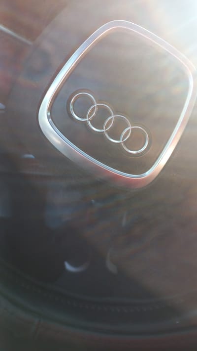 The image shows a close-up view of the Audi logo on a steering wheel. The logo consists of four interlocking rings, with a silver outline. The steering wheel itself appears to be made of black leather. Sunlight is shining on the logo, creating a bright glare and lens flare. The background is dark and blurred, suggesting the image was taken inside a car. There are no people or any other distinguishing features visible in the image. The image does not provide any information about the location in the city of Cupcini, Moldova.