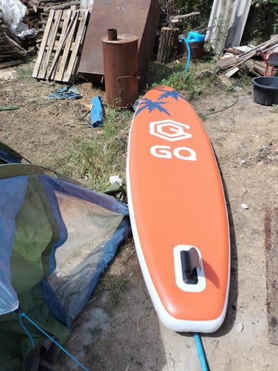 The image depicts an outdoor scene, likely a yard or a storage area, in Kudashivka, Ukraine. The foreground is dominated by an orange stand-up paddleboard with a white outline and blue palm tree graphics. The brand name "GQ" is visible in white text on the board. The paddleboard is lying on the ground, with a blue cord extending from its tail.

To the left of the paddleboard is a partially erected tent, primarily in shades of blue, green, and gray. The tent appears to be set up on dry, cracked earth with sparse dry grass.

In the background, various objects suggest a somewhat unkempt or utilitarian setting. These include a large, rusty metal cylindrical object with a pipe on top, likely a makeshift stove or kiln, and several wooden pallets stacked against a wall or fence. Further back, there are piles of wood, some corrugated roofing material, and what looks like a wooden stump with an axe embedded in it. A blue plastic watering can with a hose is also visible, suggesting recent or ongoing yard work. A black plastic bucket is in the far right.

The lighting suggests it is daytime, likely sunny given the shadows. The overall impression is one of a casual, possibly domestic, outdoor space where recreational or DIY activities might take place. There are no people visible in the image. The scene appears to be in a rural or semi-rural area.