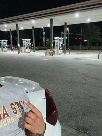 The image captures a night scene at a 7-Eleven gas station. In the foreground, a person's torso and right arm are visible, pointing with their index finger. They are wearing a grey t-shirt with maroon and gold lettering that reads "A STATE UNIVERSITY," strongly suggesting Florida State University branding. A beaded bracelet is worn on their right wrist. The back and side of a white car are also partially visible.

The gas station canopy, adorned with the characteristic red, orange, and green stripes of 7-Eleven, is brightly lit, illuminating the multiple gas pumps beneath it, which also bear the 7-Eleven logo. One sign near a pump clearly displays "149," likely indicating a gas price. The ground is asphalt.

In the background, beyond the gas station, a road with a visible red traffic light indicates it's nighttime. A tall sign further down the road, possibly an advertisement, features a green logo on a black background, resembling a Monster Energy drink ad. The overall setting is dark, with artificial lights providing illumination, confirming the time of day. The scene is static, with no discernible activity beyond the person pointing. Based on the provided context, the location is Homosassa Springs, United States of America.