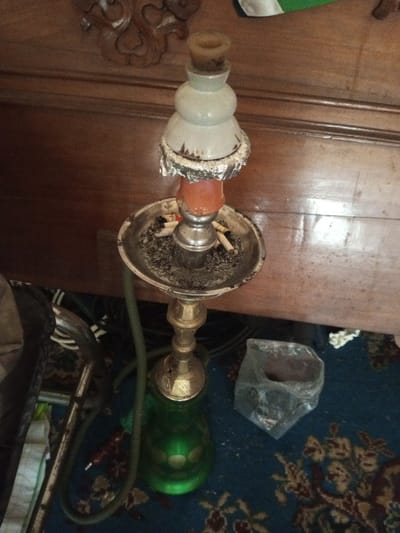A hookah with a green glass base and a gold stem rests on a carpet with a floral pattern. The hookah has a metal tray filled with ash and cigarette butts. Aluminum foil is wrapped around the top of the hookah, and a rounded white ceramic piece sits above it. A clear plastic bag sits on the carpet next to the hookah. The hookah is placed against a wooden dresser with decorative carvings.