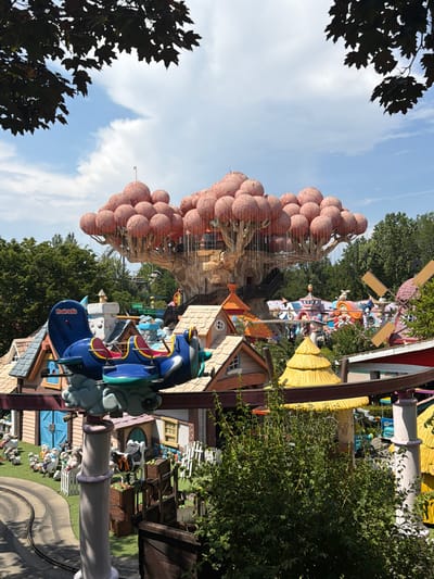 The image depicts a whimsical amusement park scene in Castelnuovo del Garda, Italy. The central feature is a large, stylized tree with a canopy of pink, balloon-like structures, resembling giant fruit. The tree is integrated with a multi-level structure, possibly a ride or themed area, accessible by wooden walkways and stairs.

In the foreground, a blue, cartoonish airplane ride vehicle is visible, suspended by a metal track. It has two rows of seats with red cushioning. The vehicle is labeled "Fruit-tella". Below the airplane, a collection of miniature houses with colorful roofs creates a storybook village atmosphere. Some of these houses have quaint details like windows and doors.

Further into the background, other amusement park attractions are scattered. A large windmill with two wooden blades stands out, painted in pastel colors. A carousel or a similar ride with colorful figures and structures is also present. The ground is covered in green grass, and a black, winding roller coaster track is partially visible. Several cartoonish figures of farm animals, including sheep and a cow, are placed along a white picket fence, adding to the playful theme.

The environment is bathed in bright daylight, with a partly cloudy sky. Lush green trees surround the park, indicating an outdoor setting. The overall impression is one of a vibrant and imaginative children's entertainment area.