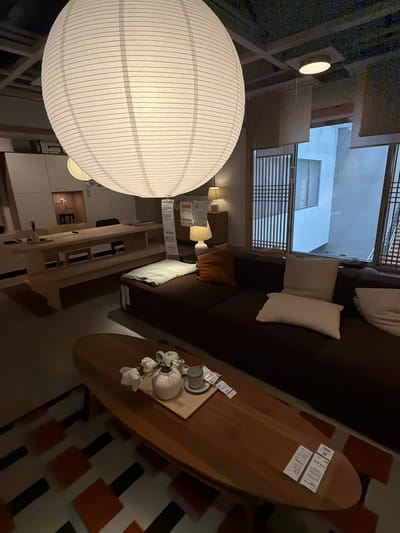 The image captures an interior scene, likely a showroom or a staged living space, within a building in Goyang-si, South Korea. Prominently featured is a large, round, white paper lantern hanging from the ceiling, casting a soft illumination. Below, a dark brown sectional sofa is arranged with several throw pillows in neutral tones. In front of the sofa, a long, oval-shaped wooden coffee table displays a small arrangement of white flowers in a white vase, a teapot, and a teacup and saucer. Several informational tags are affixed to the coffee table.
To the left of the sofa and coffee table, a long, low wooden table with shelving underneath is visible. On this table, a simple white vase and what appears to be a dark desk chair are present, suggesting a workspace or dining area. In the background, a large window with a distinctive wooden lattice design looks out onto an outdoor scene, showing a portion of a building with blue-tinted windows. The ceiling features exposed beams and a grid-like structure, with additional lighting fixtures visible. The floor is covered by a patterned rug with geometric shapes in shades of orange and black on a white background. The overall ambiance is modern and minimalist, with a focus on natural materials and soft lighting.