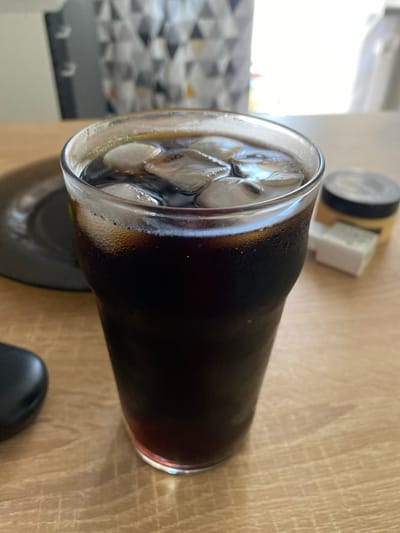 A close-up, eye-level shot captures a glass filled with dark liquid and ice cubes, set on a wooden surface. The glass appears to be sweating, indicating a cold beverage. In the background, a black plate is partially visible to the left, while to the right, a small white rectangular object and a jar with a yellow lid are out of focus. A patterned curtain and a bright window suggest an indoor setting, possibly a home or restaurant. The overall ambiance is casual and relaxed. The image does not provide any information about Maksimkovo, Russia.