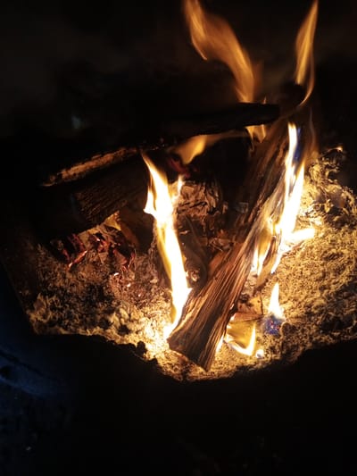 The image shows a close-up view of a bonfire. Several pieces of wood are burning, with flames and embers visible. The background is dark and out of focus, suggesting the photo was taken at night or in a dimly lit area. The ground beneath the fire appears to be covered in ash and sand. There are no people or animals visible in the image. The location in Buraly, Russia, is not discernible from the image itself.
