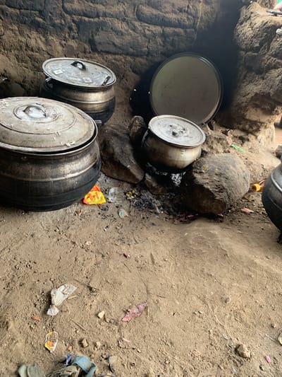 The image depicts an outdoor cooking area in what appears to be the city of Dull A, Nigeria. The scene is characterized by a rustic, earthy environment with a dirt floor and a backdrop of rough, textured walls, possibly made of mud or clay. Several large, dark metal pots are present, some with lids, indicating a cooking process is underway or has recently concluded.

In the foreground, a large pot with a lid is prominently positioned. To its left, another similar pot is visible, also with its lid. In the background, a large, flat, circular metal object, possibly a tray or a lid, rests against a rocky formation. Adjacent to the cooking pots, there are several large rocks, which may be used to support the cooking vessels or as part of the hearth. Scattered on the ground around the pots are remnants of food, ash, and debris, including small pieces of plastic and paper packaging, suggesting a meal preparation area.

The overall impression is one of a traditional, perhaps communal, cooking setup in an open-air environment. The lighting suggests it is daytime, with some shadows indicating the direction of light. There are no people visible in the image.