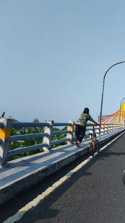 A person is standing with a bicycle on the sidewalk of a bridge. The person is wearing a camouflage-patterned shirt and dark pants, and their right hand is resting on the handlebars of the bicycle. The bicycle is orange and black and is positioned to the right of the person. The bridge has a concrete railing with metal bars, and yellow markings on the pillars. Beyond the railing, there are green banana leaves and some buildings visible. The bridge's architecture features colorful, arched decorative elements in yellow and orange. The roadway is paved and has a broken yellow line. The sky is clear and blue. The scene appears to be in Trosobo, Indonesia, given the surrounding vegetation and architectural style.